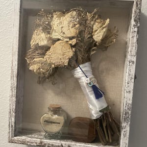 May include: A framed display featuring a dried flower bouquet with a white ribbon and blue tassel. A small glass bottle with the word "Honeymoon" and a wooden block are also in the frame. The frame is white with a distressed finish.