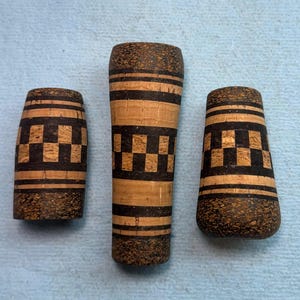 Puede incluir: Tres empuñaduras cilíndricas para cañas de pescar hechas de corcho natural con detalles de corcho compuesto marrón oscuro. Cada pieza presenta un patrón de tablero de ajedrez decorativo y bandas horizontales de material oscuro en el corcho.
