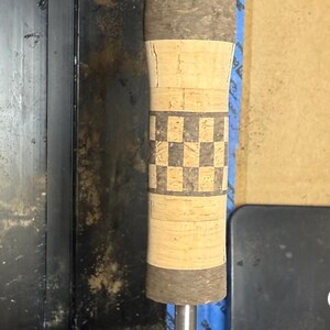 Puede incluir: Un mango de caña de pescar personalizado con un agarre de corcho y un patrón de tablero de ajedrez decorativo. El mango está montado sobre una varilla de metal, con el logotipo "CRB Custom Rod Builder" visible en el fondo.
