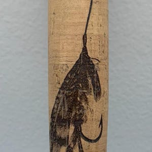 Peut inclure: Une poignée de canne à pêche en liège avec un motif de mouche noir et blanc brûlé à la surface.