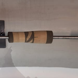 Puede incluir: Una caña de pescar con mango de corcho y un eje plateado. El mango tiene un patrón marrón y negro. La caña está montada sobre un soporte negro con un logotipo blanco y verde. La caña está diseñada para la pesca.