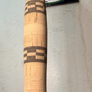 Puede incluir: Primer plano de un mango de caña de pescar hecho de corcho con bandas decorativas de color marrón oscuro. El mango está unido a una varilla de metal plateado. La imagen incluye una pegatina que dice "Rods of Indiana".