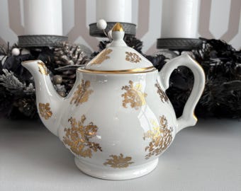 May include: White porcelain teapot with gold floral designs and accents. The teapot has a curved handle and a lid with a gold finial. The teapot is sitting on a white surface.