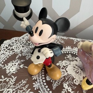 May include: A Mickey Mouse figurine in a classic outfit, holding a black top hat. The figure has black ears, a black body, and tan shoes. The background is a patterned wall and a brown surface with a white lace doily.