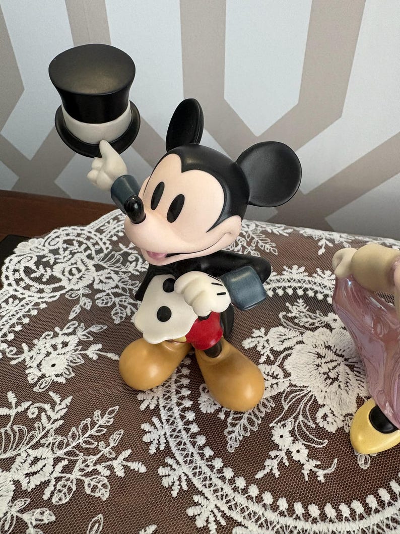 May include: A Mickey Mouse figurine in a classic outfit, holding a black top hat. The figure has black ears, a black body, and tan shoes. The background is a patterned wall and a brown surface with a white lace doily.