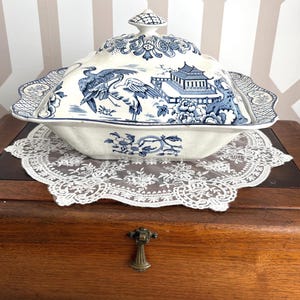 Op de afbeelding: Een crèmekleurige serveerschaal met deksel, versierd met een blauw-wit Aziatisch geïnspireerd ontwerp. De rechthoekige schaal heeft geschulpte randen en een decoratieve knop. Het staat op een wit kanten kleedje.