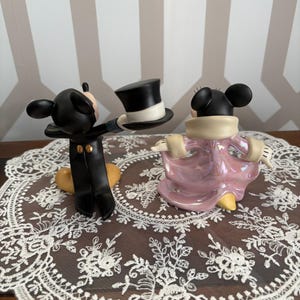 May include: Two Disney Mickey and Minnie Mouse figurines. Mickey is in a black tuxedo holding a top hat. Minnie is in a pink dress with a cream-colored collar. The figurines are on a white lace doily.