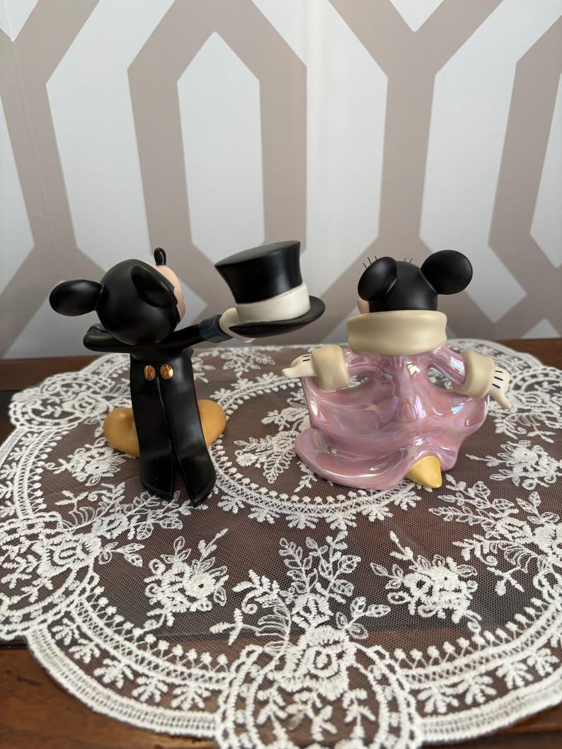 May include: Two Disney Mickey and Minnie Mouse figurines. Mickey is in a black tuxedo holding a top hat. Minnie is in a pink dress with a cream-colored collar. The figurines are on a white lace doily.