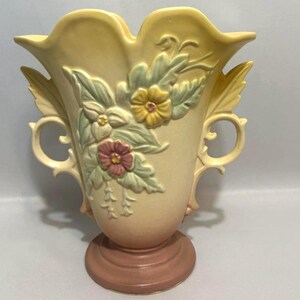 May include: A vintage ceramic vase in a soft yellow hue, featuring embossed floral designs in shades of pink, yellow, and green. The vase has a flared top, two side handles, and a pedestal base, creating an elegant, decorative piece.