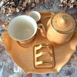 Pode incluir: Um conjunto de chá vintage cor de pêssego com detalhes dourados, incluindo uma chaleira, uma chávena, um pires e um pequeno jarro de creme, dispostos numa bandeja a condizer. O conjunto tem um design clássico e elegante. A bandeja inclui um cinzeiro.