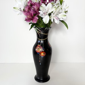 Puede incluir: Jarrón de cerámica negro con borde dorado y un diseño de linternas rojas y naranjas colgando de una cuerda dorada. El jarrón está lleno de flores blancas y moradas.