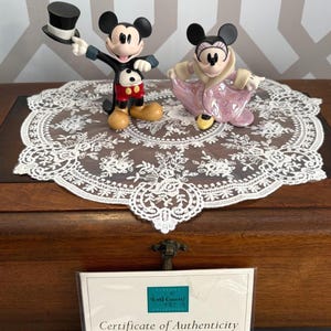 May include: Two Disney figurines of Mickey and Minnie Mouse. Mickey is wearing a black tuxedo with a top hat, red shorts, and yellow shoes. Minnie is in a pink dress and shoes. They are on a white lace doily.