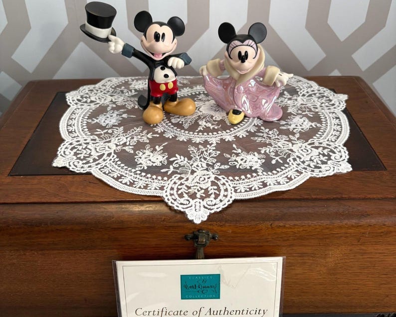 May include: Two Disney figurines of Mickey and Minnie Mouse. Mickey is wearing a black tuxedo with a top hat, red shorts, and yellow shoes. Minnie is in a pink dress and shoes. They are on a white lace doily.