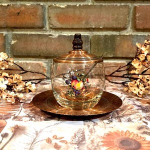 Puede incluir: Un tarro de cristal transparente con tapa y base color cobre, con un diseño floral y de frutas. El tarro incluye una cucharita y se apoya en un plato a juego color cobre. El fondo es una pared de ladrillos y un arreglo floral.
