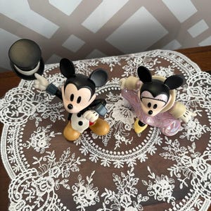 May include: Two Mickey Mouse figurines. One Mickey is dressed in a tuxedo and holding a black top hat. The other Mickey is wearing a pink outfit. Both figurines have black ears and yellow shoes. They are displayed on a white lace doily.