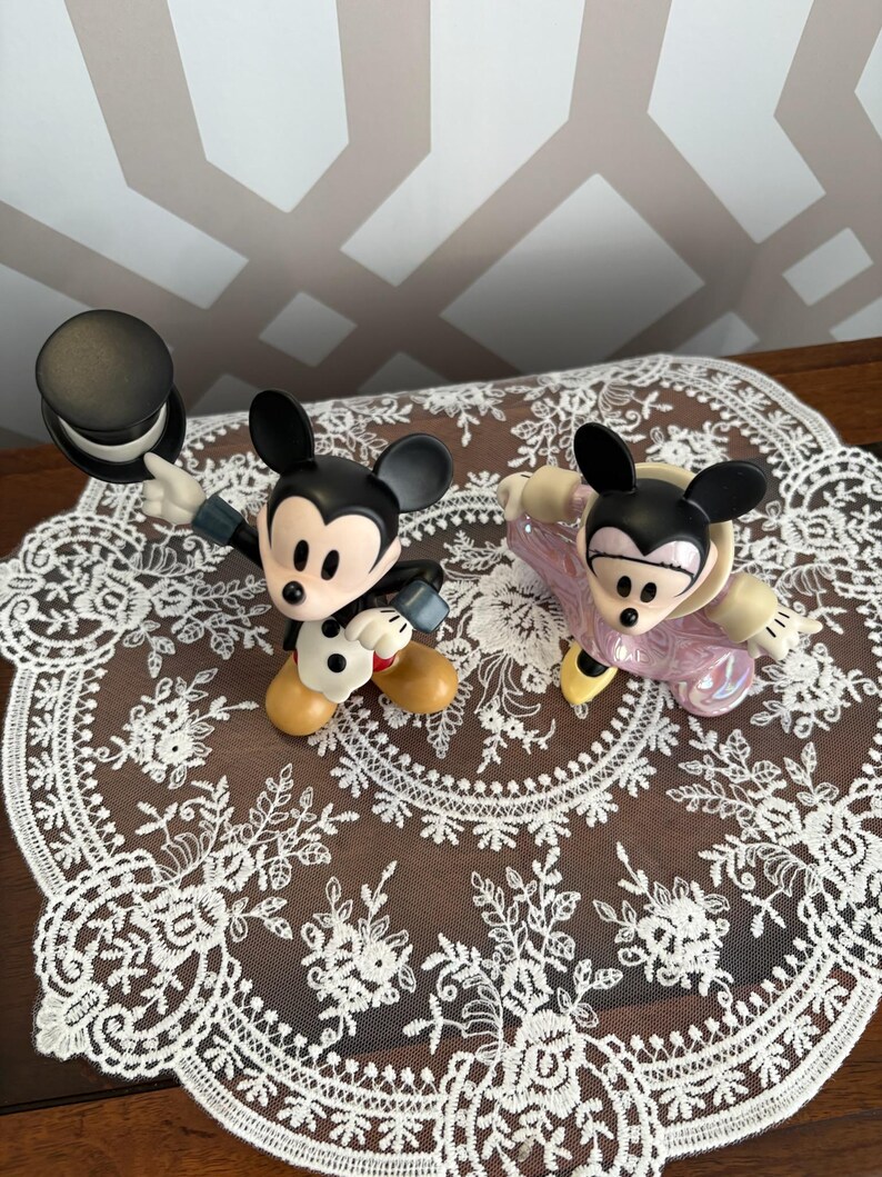 May include: Two Mickey Mouse figurines. One Mickey is dressed in a tuxedo and holding a black top hat. The other Mickey is wearing a pink outfit. Both figurines have black ears and yellow shoes. They are displayed on a white lace doily.