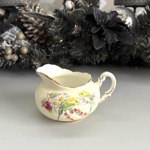 Puede incluir: Una pequeña jarra de crema de cerámica de color crema con borde y asa dorados. La jarra de crema está decorada con un diseño floral y de pájaros con periquitos verdes, flores rosas y margaritas blancas. Es una pieza decorativa.