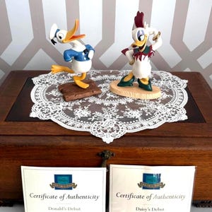 May include: Two Disney character figurines, Donald Duck and Daisy Duck, are displayed on a wooden box with a white lace doily. Donald Duck wears a sailor outfit, and Daisy Duck is in a red and white outfit. Certificates of Authenticity are visible.