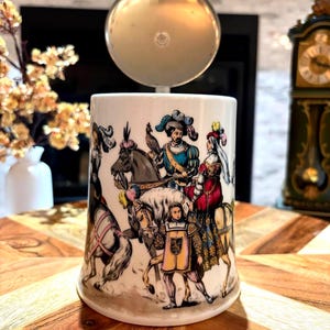 May include: A white ceramic beer stein featuring a colorful illustration of historical figures on horseback and on foot, wearing elaborate period clothing and feathered hats. The stein is topped with a circular, polished silver-colored metal lid.