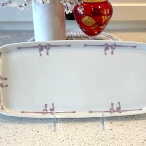 Puede incluir: Una fuente rectangular blanca con esquinas redondeadas, adornada con un diseño de lazo lavanda repetido a lo largo de los bordes. La fuente se exhibe sobre un soporte de metal transparente, adecuada para servir aperitivos o postres.