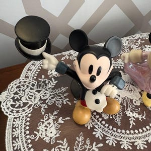 May include: A Mickey Mouse figurine in a classic pose, wearing a black tuxedo and gloves, with a black top hat. The figurine has a tan face and feet. The background is a brown lace doily.