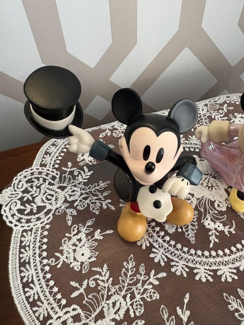 May include: A Mickey Mouse figurine in a classic pose, wearing a black tuxedo and gloves, with a black top hat. The figurine has a tan face and feet. The background is a brown lace doily.