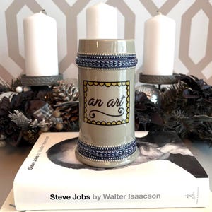 May include: A beige ceramic beer stein with blue and white decorative bands. The stein has a yellow-bordered label with the words "an art" in black script. The stein is sitting on a book titled "Steve Jobs by Walter Isaacson".