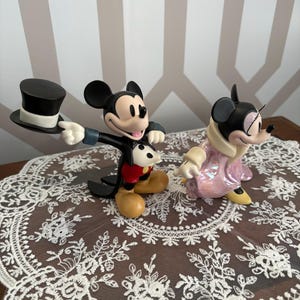 May include: Two Disney figurines of Mickey and Minnie Mouse. Mickey is holding a black top hat, wearing a black suit, red shorts, and yellow shoes. Minnie is in a pink coat and yellow shoes. They are on a white lace doily.