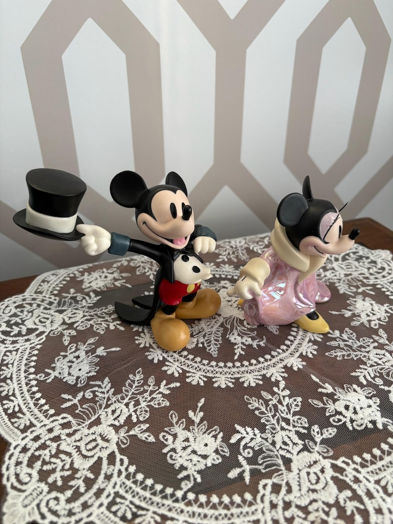 May include: Two Disney figurines of Mickey and Minnie Mouse. Mickey is holding a black top hat, wearing a black suit, red shorts, and yellow shoes. Minnie is in a pink coat and yellow shoes. They are on a white lace doily.