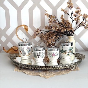 May include: Four decorative egg cups with silver lids and handles, each with a unique floral or animal design. The cups sit on a mirrored tray with ornate edges, alongside a glass swan and a vase of flowers.