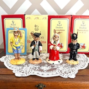 May include: Four Royal Doulton Brambly Hedge Bunnykins figurines in various outfits, including a tourist, a gentleman in a suit, a woman in a red dress, and a police officer. The figurines are displayed in front of their original boxes.