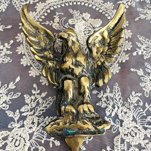May include: A vintage brass eagle sculpture with outstretched wings. The eagle is detailed with textured feathers and a weathered finish. The sculpture has a loop at the bottom for hanging. The background is a white lace fabric.
