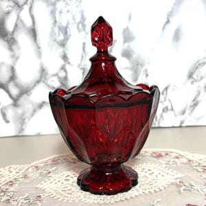 Puede incluir: Un cuenco de cristal rojo intenso con tapa a juego, que presenta un patrón facetado similar a un diamante en el exterior. El recipiente se apoya sobre una base circular y está rematado con un pomo geométrico puntiagudo en la tapa extraíble.