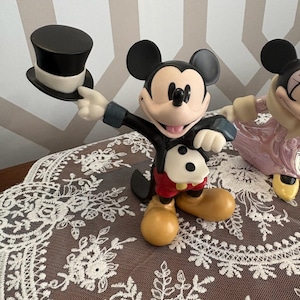 May include: A Mickey Mouse figurine holding a black top hat. The figure is wearing a black tuxedo jacket, red shorts, and yellow shoes. The figurine is on a brown surface with a white lace overlay.
