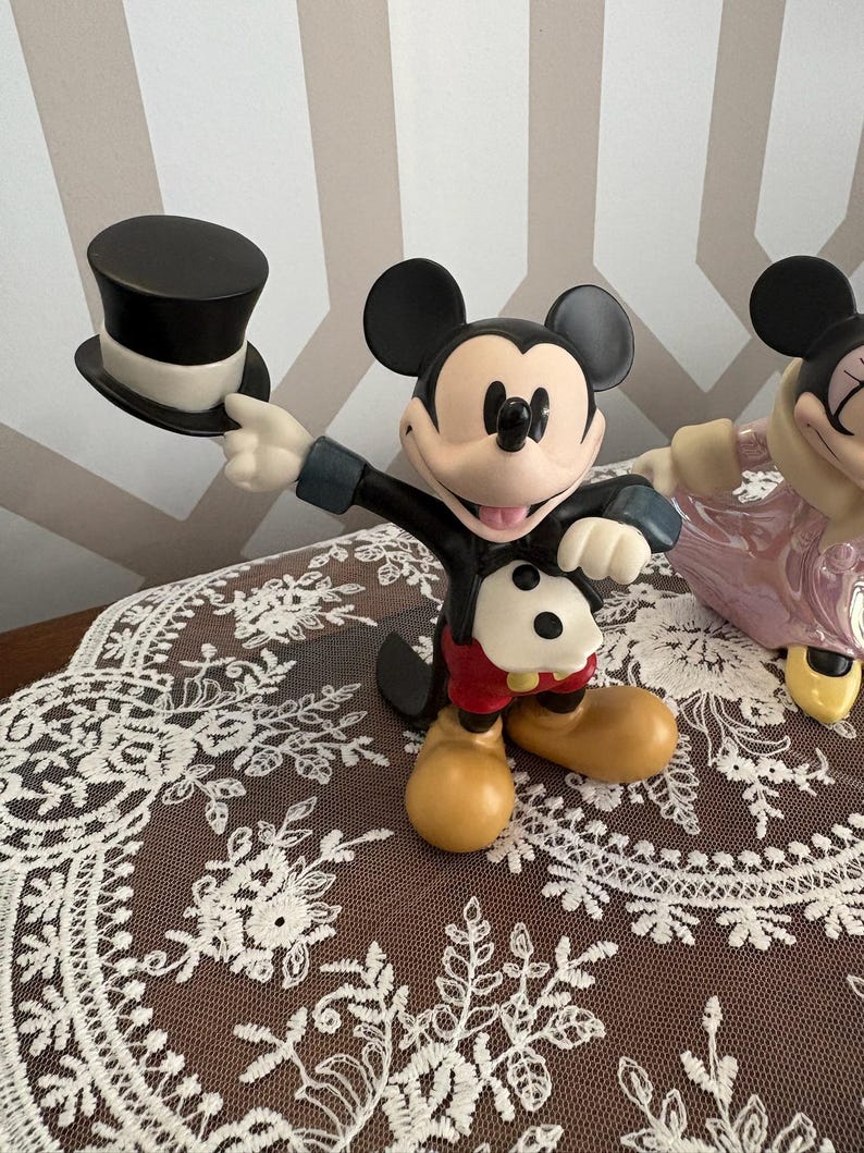 May include: A Mickey Mouse figurine holding a black top hat. The figure is wearing a black tuxedo jacket, red shorts, and yellow shoes. The figurine is on a brown surface with a white lace overlay.