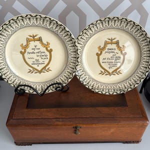 Puede incluir: Dos platos decorativos con bordes ornamentados y texto. Los platos son de color crema con un diseño dorado y marrón. Uno de los platos dice "If any one speaks evil you, let your life be such that no one will believe him."