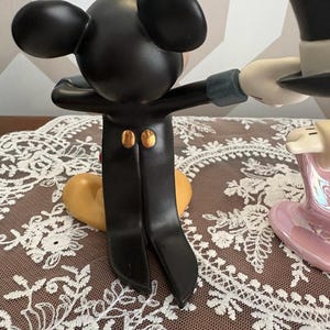 May include: A black Mickey Mouse figurine in a tuxedo with gold buttons, arms outstretched. The figurine is interacting with a white-gloved hand holding a black top hat. A heart-shaped base is visible.