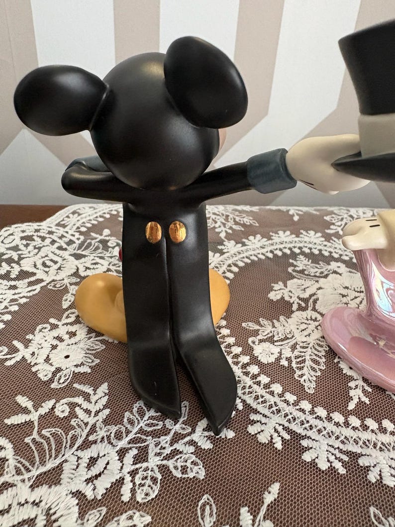 May include: A black Mickey Mouse figurine in a tuxedo with gold buttons, arms outstretched. The figurine is interacting with a white-gloved hand holding a black top hat. A heart-shaped base is visible.