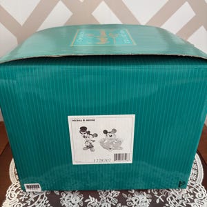 May include: A teal box with vertical lines, featuring a label with Mickey & Minnie illustrations. The box is on a table with a white lace doily. The top of the box has the words "Classics Collection".