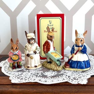 May include: Four ceramic rabbit figurines in different outfits, including a chef, a farmer with a wheelbarrow, and a woman with a broom. The figurines are on a white lace doily, with a red box in the background.