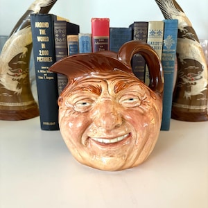 Puede incluir: Una jarra de cerámica marrón con un diseño de cara sonriente. La jarra tiene un asa y un pico. La cara está detallada con ojos, nariz y una amplia sonrisa. Libros y cuernos decorativos en el fondo.