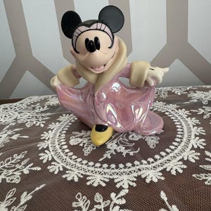 May include: A Minnie Mouse figurine in a pink iridescent dress and coat, with black ears and a yellow shoe. The figurine is posed with one arm extended. The figurine is on a white lace tablecloth.