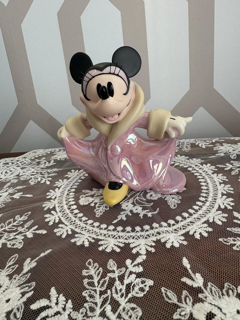 May include: A Minnie Mouse figurine in a pink iridescent dress and coat, with black ears and a yellow shoe. The figurine is posed with one arm extended. The figurine is on a white lace tablecloth.