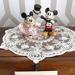 May include: Two Disney figurines of Mickey and Minnie Mouse. Mickey is in a black suit with red shorts, holding a top hat. Minnie is in a pink dress. They are on a white lace doily, set on a wooden surface.
