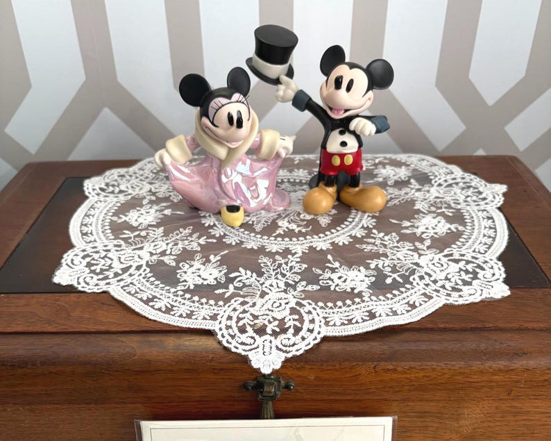 May include: Two Disney figurines of Mickey and Minnie Mouse. Mickey is in a black suit with red shorts, holding a top hat. Minnie is in a pink dress. They are on a white lace doily, set on a wooden surface.