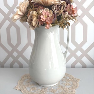 Könnte beinhalten: Eine weiße Keramikkrug mit Henkel, gefüllt mit einem Strauß Kunstblumen in Rosa, Beige und Burgunder. Der Krug steht auf einer beigen Spitzendecke. Der Hintergrund hat ein geometrisches Muster.
