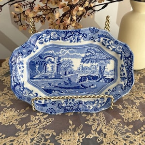 Peut inclure: Assiette décorative en céramique bleue et blanche avec une scène de paysage. L'assiette a un bord festonné et est présentée sur un présentoir en métal doré. L'assiette présente une illustration détaillée d'une scène rurale.