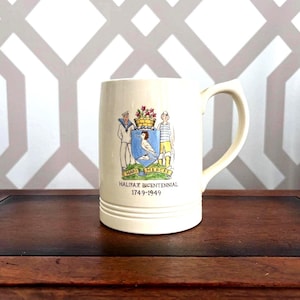 May include: Cream-colored ceramic mug with a curved handle. The mug features a colorful illustration of two figures, a coat of arms, and the text "HALIFAX BICENTENNIAL 1749-1949". The mug is set on a dark wooden surface.