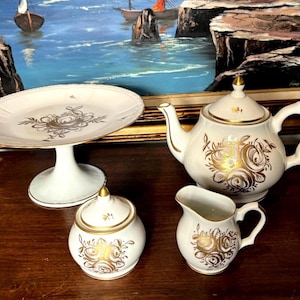 May include: A vintage white porcelain tea set with gold floral patterns. The set includes a teapot, creamer, sugar bowl, and a cake stand. The teapot has a lid with a gold finial. The cake stand has a pedestal base.