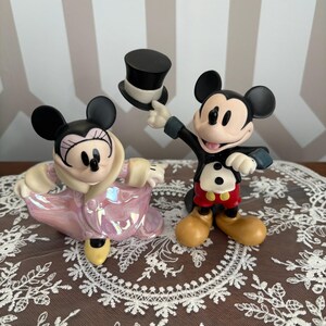 May include: Two ceramic figurines of Mickey and Minnie Mouse. Mickey is in a tuxedo, holding a black top hat. Minnie is wearing a pink iridescent coat. Both have black ears and are smiling.
